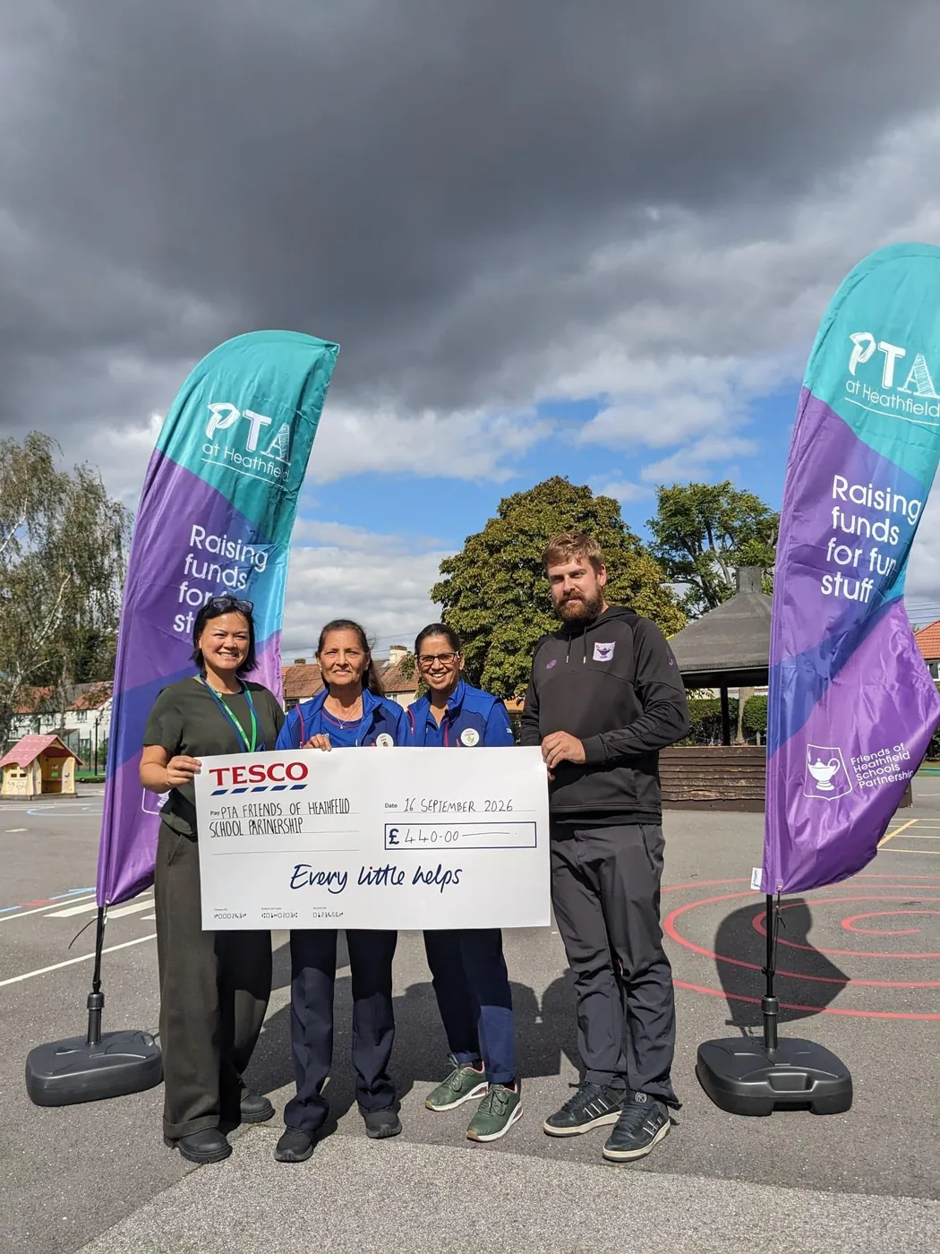 Four adults standing in a school playground holding a large Tesco cheque for £440, between two PTA flag banners reading Raising funds for fun stuff